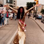 A person in a gold dress marches in a Pride parade. Their arms are raised high in celebration.