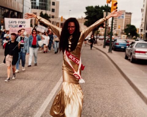 A person in a gold dress marches in a Pride parade. Their arms are raised high in celebration.