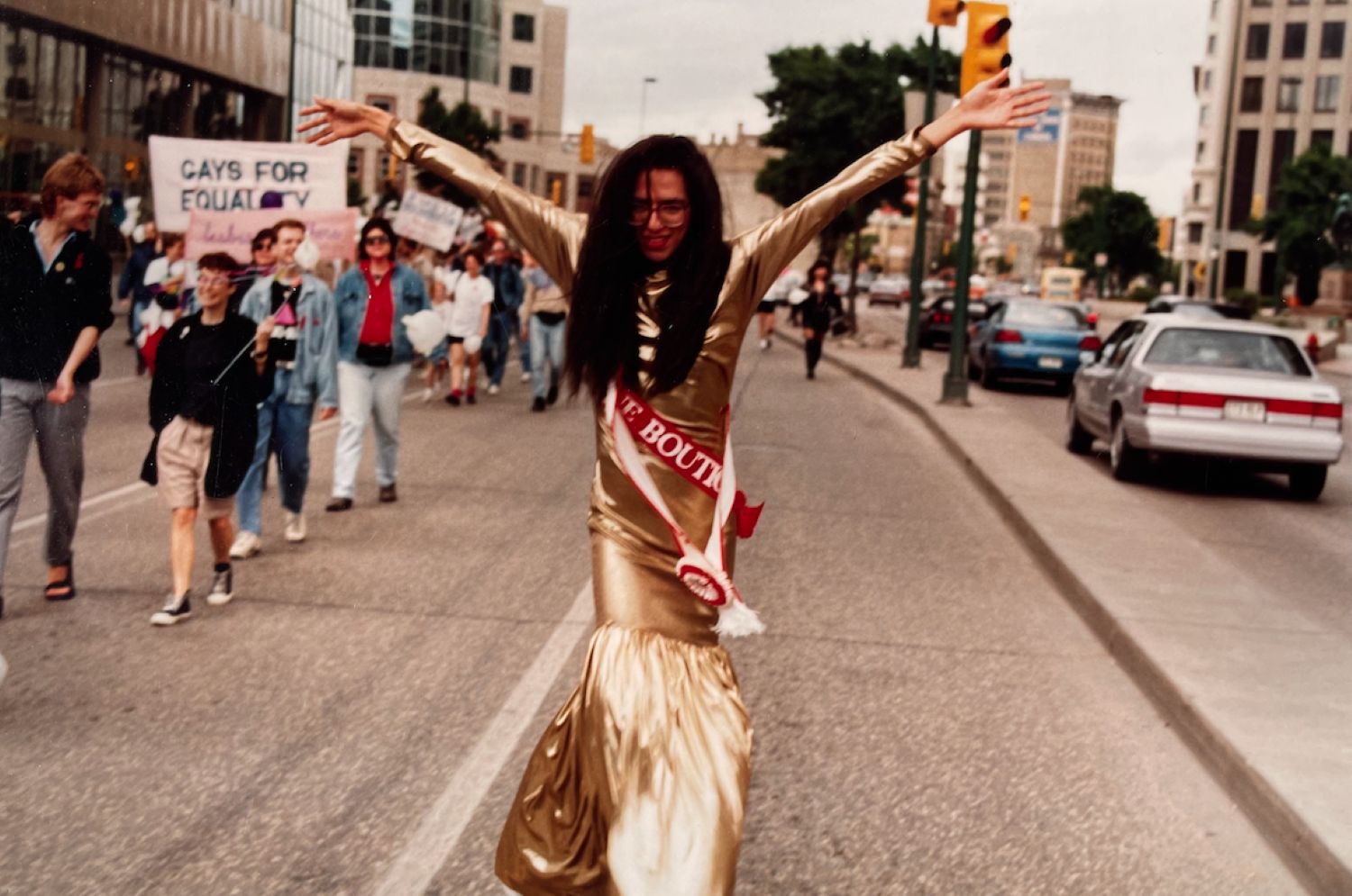 A person in a gold dress marches in a Pride parade. Their arms are raised high in celebration.