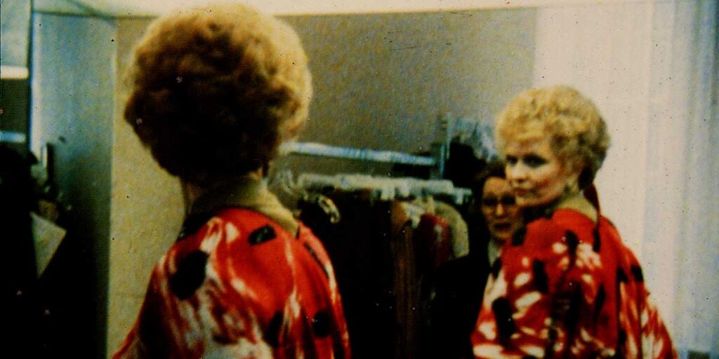 A woman tries on an outfit while looking at herselfin a mirror. She is wearing a red garment patterned with white and black. A sales woman can be seen in the mirror, standing behind her.