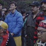 An archival video image shows a group of Indigenous land defenders standing together in protest. Elders in regalia are seated in the front row, while people in street clothes stand behind them. A lush forest is in the background.