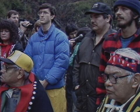 An archival video image shows a group of Indigenous land defenders standing together in protest. Elders in regalia are seated in the front row, while people in street clothes stand behind them. A lush forest is in the background.