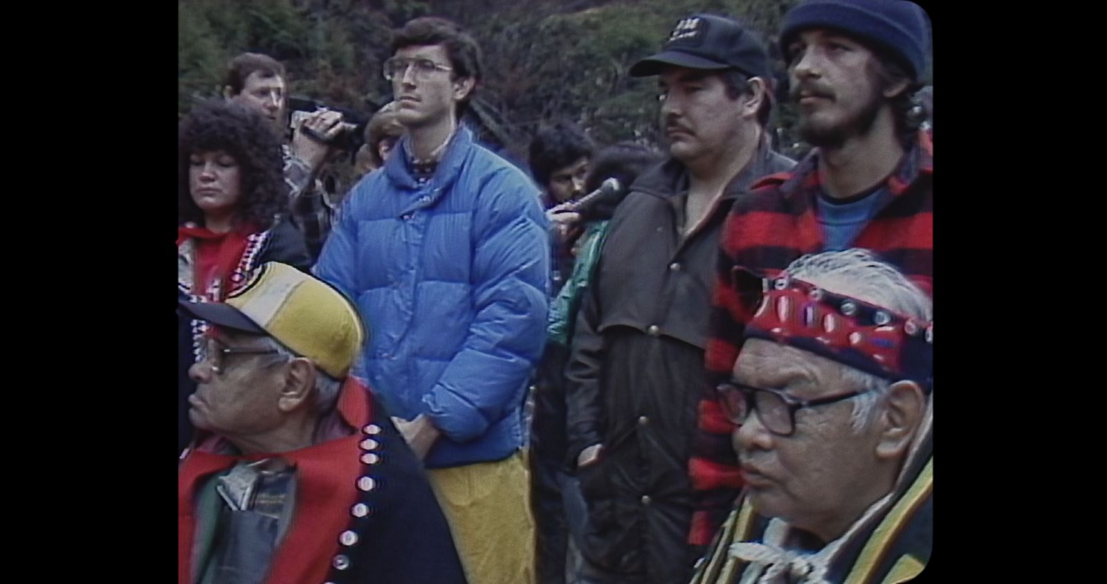 An archival video image shows a group of Indigenous land defenders standing together in protest. Elders in regalia are seated in the front row, while people in street clothes stand behind them. A lush forest is in the background.