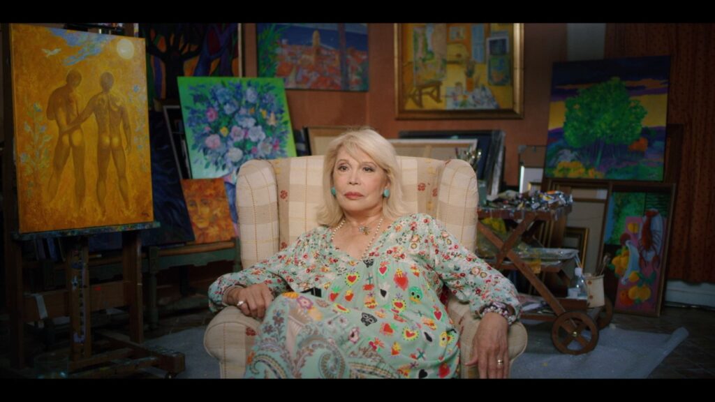 April Lear is a mature blonde woman seated in a light blue dress patterned with dots of different sizes an colours. She is surrounded by paintings, including one of two nude men on screen left.