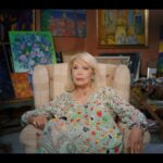April Lear is a mature blonde woman seated in a light blue dress patterned with dots of different sizes an colours. She is surrounded by paintings, including one of two nude men on screen left.