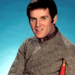 Actor Charles Grodin appears in an archival photo from the 1970s. He is a white man with brown hair and brown eyes, wearing a dark grey sweater. He is seated on a red chair with a blue background behind him.