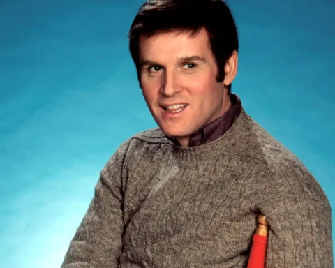 Actor Charles Grodin appears in an archival photo from the 1970s. He is a white man with brown hair and brown eyes, wearing a dark grey sweater. He is seated on a red chair with a blue background behind him.