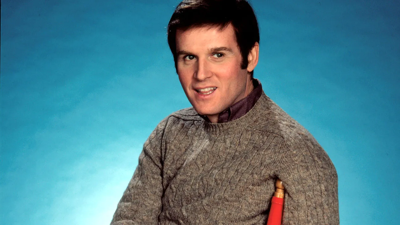 Actor Charles Grodin appears in an archival photo from the 1970s. He is a white man with brown hair and brown eyes, wearing a dark grey sweater. He is seated on a red chair with a blue background behind him.