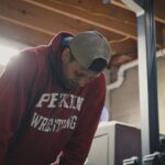 A middle aged male athlete stands in a gym with his hands on his knees and his head down. He seems anguished, and is wearing a burgundy sweatshirt with Peking Wrestling in white letters.