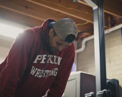 A middle aged male athlete stands in a gym with his hands on his knees and his head down. He seems anguished, and is wearing a burgundy sweatshirt with Peking Wrestling in white letters.
