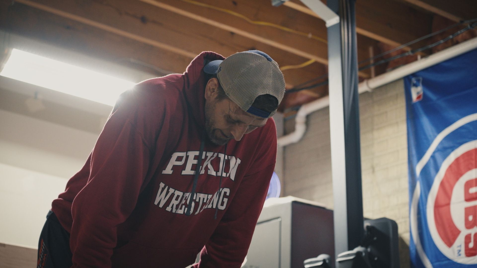 A middle aged male athlete stands in a gym with his hands on his knees and his head down. He seems anguished, and is wearing a burgundy sweatshirt with Peking Wrestling in white letters.