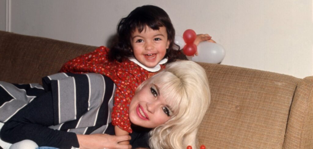 A vintage photo shows Jayne Mansfield, a white woman with blonde hair and brown eyes, lying down on a tan plaid couch. Her young daughter is on her shoulders. She is wearing a red dress, and has brown hair and brown eyes. They are both smiling.