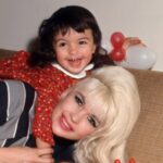 A vintage photo shows Jayne Mansfield, a white woman with blonde hair and brown eyes, lying down on a tan plaid couch. Her young daughter is on her shoulders. She is wearing a red dress, and has brown hair and brown eyes. They are both smiling.