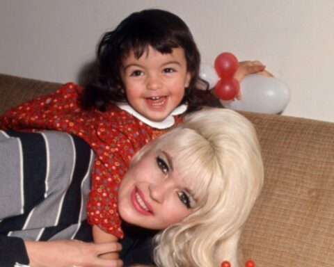 A vintage photo shows Jayne Mansfield, a white woman with blonde hair and brown eyes, lying down on a tan plaid couch. Her young daughter is on her shoulders. She is wearing a red dress, and has brown hair and brown eyes. They are both smiling.