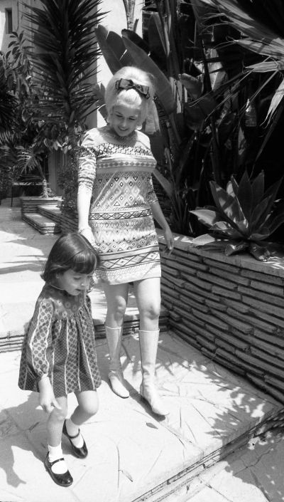A black and white photo of Jayne Mansfield walking with her young daughter. They are both wearing patterned dresses and are holding hands while walking down stone steps in a garden.