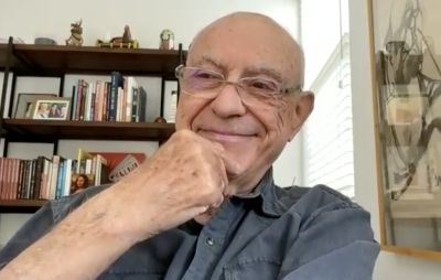 Actor Alan Arkin appears on a Zoom screen. He is an elderly white man who is bald and wearing glasses. He has his hand to his chin and a big smile on his face.