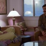 A woman in her late 20s sits cross-legged on a couch inside her apartment. A man of a similar age sits on a chair opposite her as she interviews him about becoming her roommate.