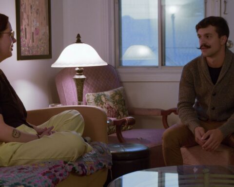 A woman in her late 20s sits cross-legged on a couch inside her apartment. A man of a similar age sits on a chair opposite her as she interviews him about becoming her roommate.