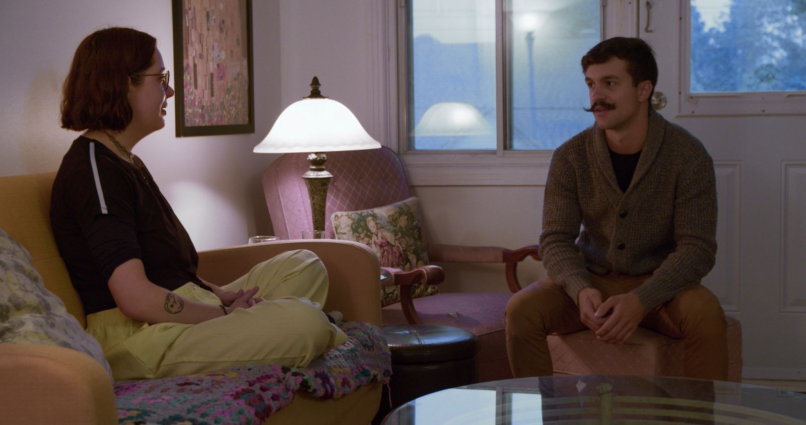 A woman in her late 20s sits cross-legged on a couch inside her apartment. A man of a similar age sits on a chair opposite her as she interviews him about becoming her roommate.