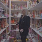 A woman in her 60s, white with blonde hair, stands in a closet. She is surrounded by shelves full of Barbie dolls in various styles and outfits.