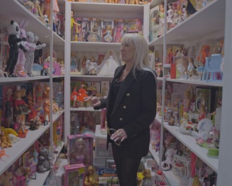A woman in her 60s, white with blonde hair, stands in a closet. She is surrounded by shelves full of Barbie dolls in various styles and outfits.