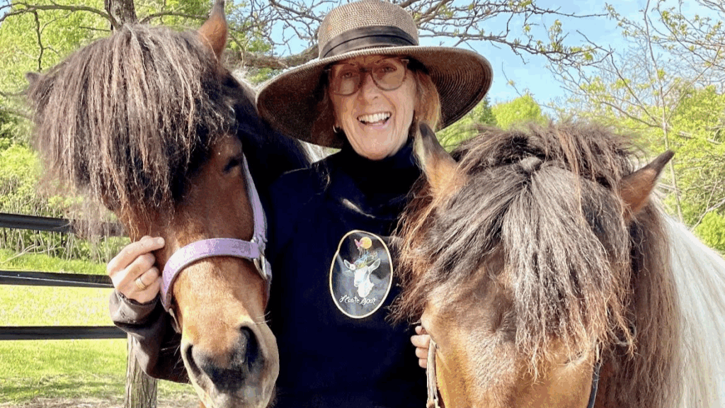 A woman in her sixties is smiling and standing with two brown horses. She is wearing a broad-rimmed hat, glasses, and a black sweatshirt with a goat logo in the centre that says Haute Goat.