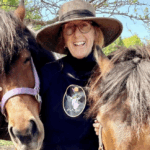 A woman in her sixties is smiling and standing with two brown horses. She is wearing a broad-rimmed hat, glasses, and a black sweatshirt with a goat logo in the centre that says Haute Goat.