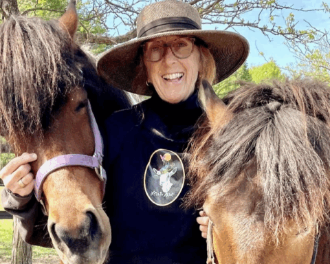 A woman in her sixties is smiling and standing with two brown horses. She is wearing a broad-rimmed hat, glasses, and a black sweatshirt with a goat logo in the centre that says Haute Goat.