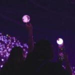Fans hold up lights at a BTS concert. The view shows a crowd of young people bathed in purple light.