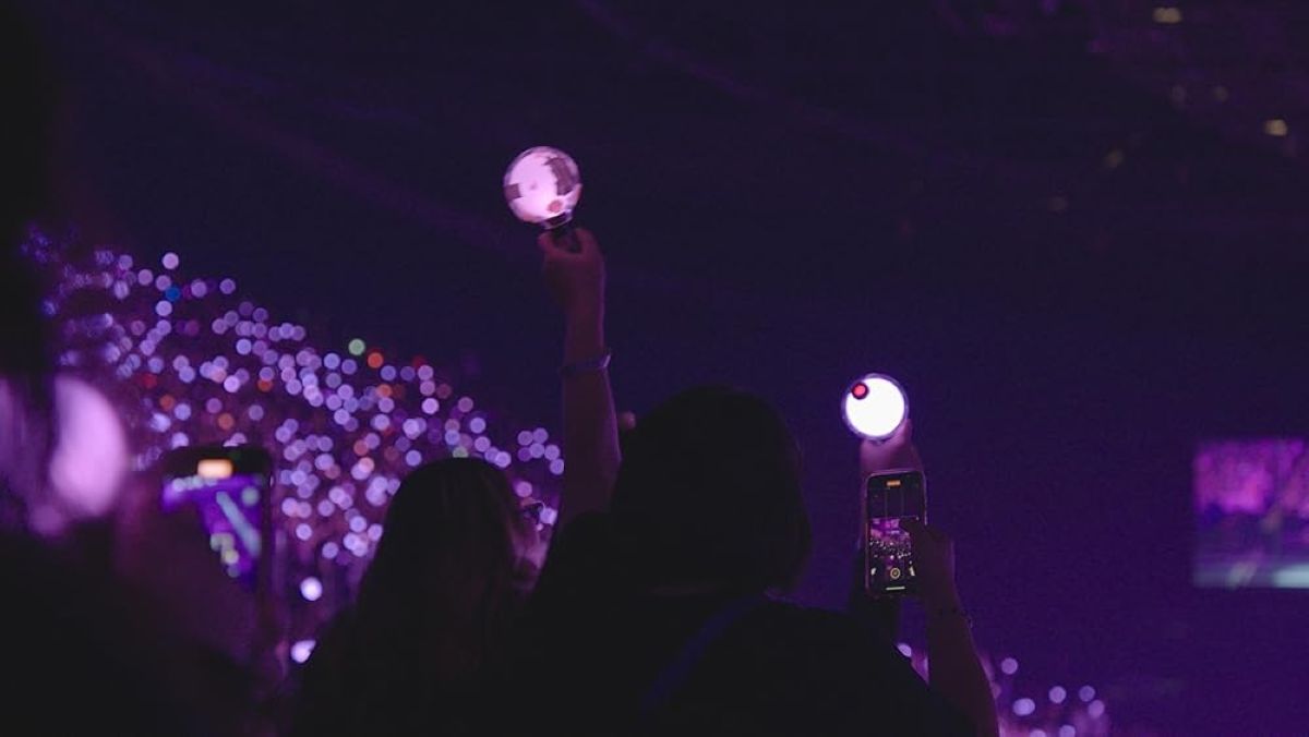 Fans hold up lights at a BTS concert. The view shows a crowd of young people bathed in purple light.