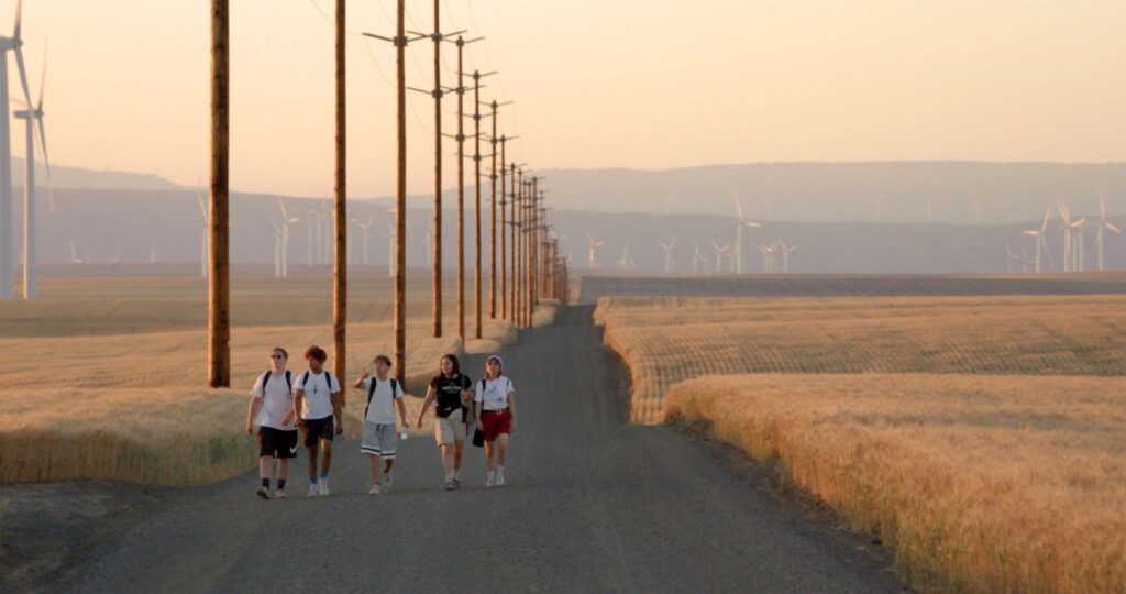 A still from Gasoline Rainbow showing people walking on an empty highway with yellow fields on either side.