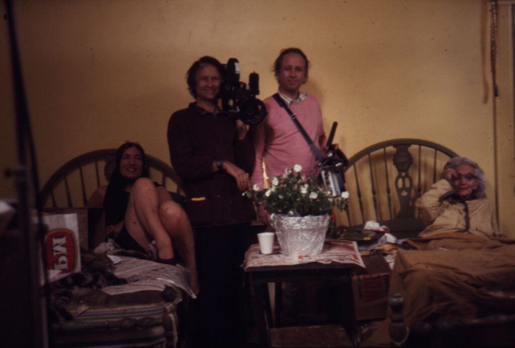 A behind the scenes still from the production of Grey Gardens during the 1970s. The Maysles brothers are in the centre holding a camera and microphone. They are standing between two beds: Little Edie is seated on the left bed and Big Edie is lying down on the right.