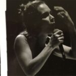 A black and white image with scratched film strip framing. It shows a woman in a tank top singing. She is holding a microphone in one hand with the other hand raised in the air.