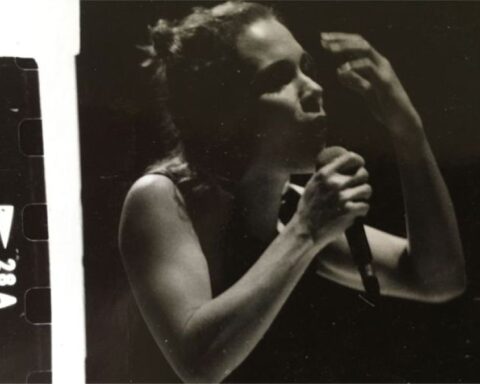 A black and white image with scratched film strip framing. It shows a woman in a tank top singing. She is holding a microphone in one hand with the other hand raised in the air.