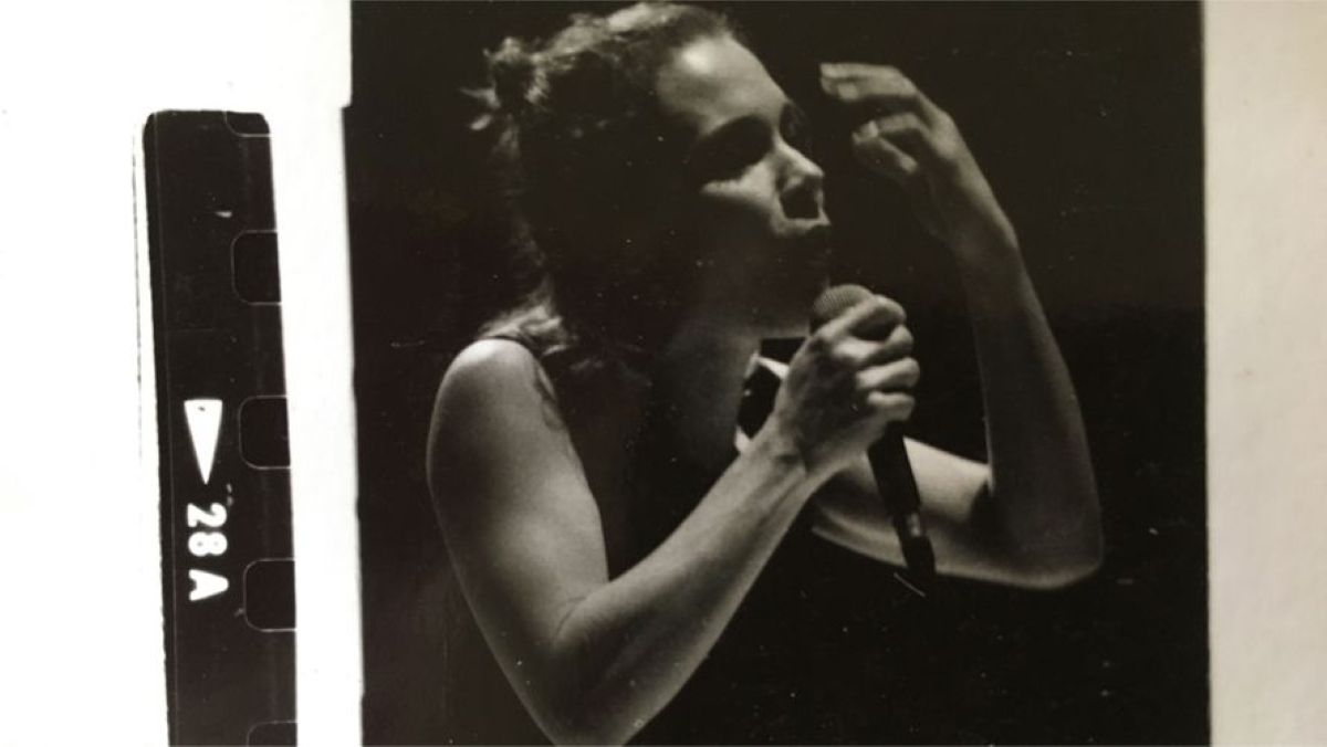 A black and white image with scratched film strip framing. It shows a woman in a tank top singing. She is holding a microphone in one hand with the other hand raised in the air.