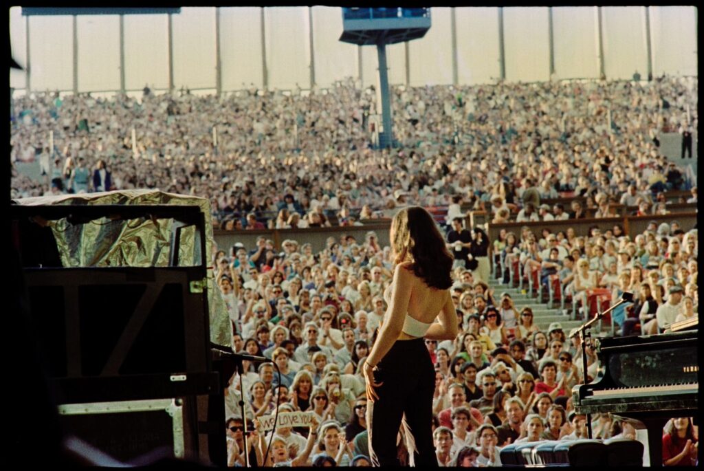 An archival photo of singer Paula Cole at Lilith Fair. She is on stage, viewed from behind, and the photo shows her looking out at the crowd of people.