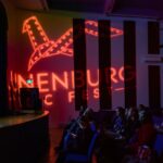 A speaker addresses an audience from a stage in a darkened theatre. The screen advertises a pitch competition and the logo for Lunenburg Doc Fest appears in lights on the theatre wall.