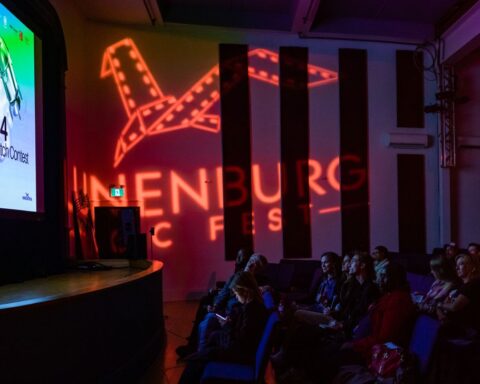 A speaker addresses an audience from a stage in a darkened theatre. The screen advertises a pitch competition and the logo for Lunenburg Doc Fest appears in lights on the theatre wall.