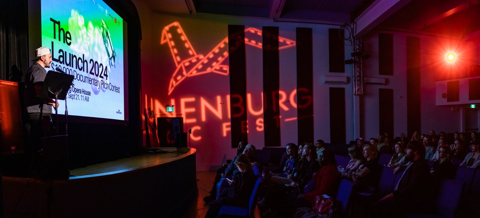 A speaker addresses an audience from a stage in a darkened theatre. The screen advertises a pitch competition and the logo for Lunenburg Doc Fest appears in lights on the theatre wall.