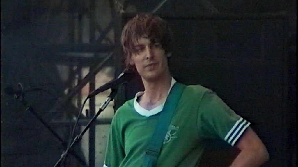 An archival image of Pavement's Stephen Malkmus on stage at Lollapalooza 1995. He is wearing a green t-shirt with a white-ringed collar and standing at a microphone.