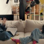 Two women sit at opposite ends of a couch. They are facing each other and having a conversation in American Sign Language.