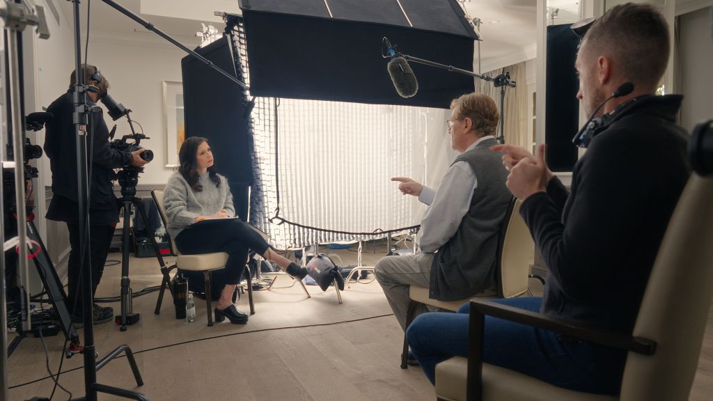 A behind the scenes image of filmmaker Shoshannah Stern conducting an interview with writer/director Aaron Sorkin. They are surrounded by cameras, lights, and production crew.