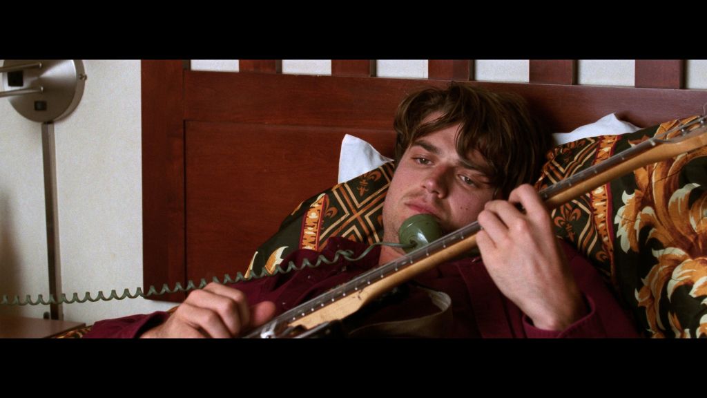 A 1990s rocker strums on his guitar in bed. The wall behind him is maroon-red.