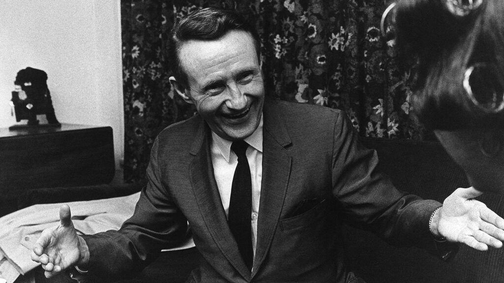 A black and white image of a man in a suit during the 1960s. He is white with brown hair. He is smiling and has his hands held out making a positive gesture.
