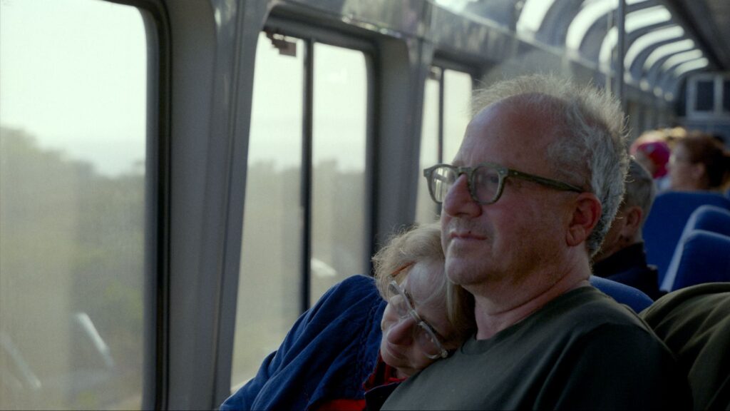 A husband and wife sit together on a bus. She is resting her head on his shoulder and he looks out the window. They are both white and wearing glasses and in their early 60s.