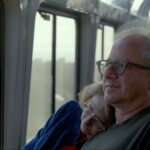 A husband and wife sit together on a bus. She is resting her head on his shoulder and he looks out the window. They are both white and wearing glasses and in their early 60s.