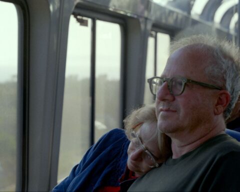 A husband and wife sit together on a bus. She is resting her head on his shoulder and he looks out the window. They are both white and wearing glasses and in their early 60s.