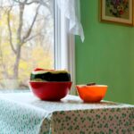 A partially cut watermelon rests in a red bowl on a table. There is a smaller orange bowl beside it. The table has a white tablecloth with a holly berry style pattern, and is positioned near an open window through which the sun shines on the melon.