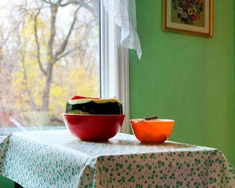A partially cut watermelon rests in a red bowl on a table. There is a smaller orange bowl beside it. The table has a white tablecloth with a holly berry style pattern, and is positioned near an open window through which the sun shines on the melon.