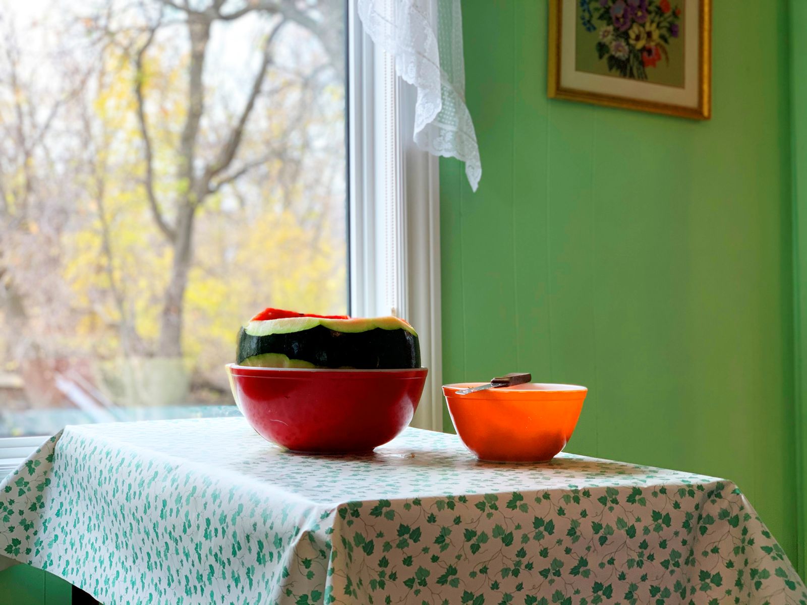 A partially cut watermelon rests in a red bowl on a table. There is a smaller orange bowl beside it. The table has a white tablecloth with a holly berry style pattern, and is positioned near an open window through which the sun shines on the melon.